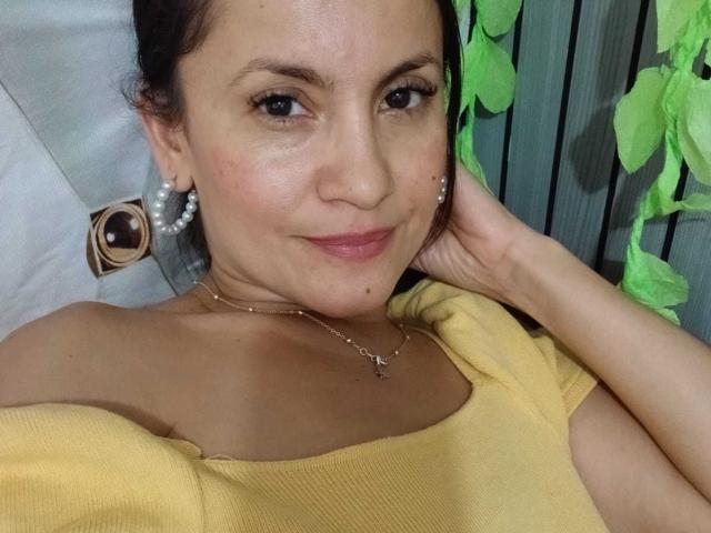 SolaraDubois, con una sonrisa encantadora, ojos marrones, y vistiendo una blusa amarilla con un hombro descubierto, mostrando sus senos y un collar pequeño, con decoraciones de hojas verdes en el fondo..