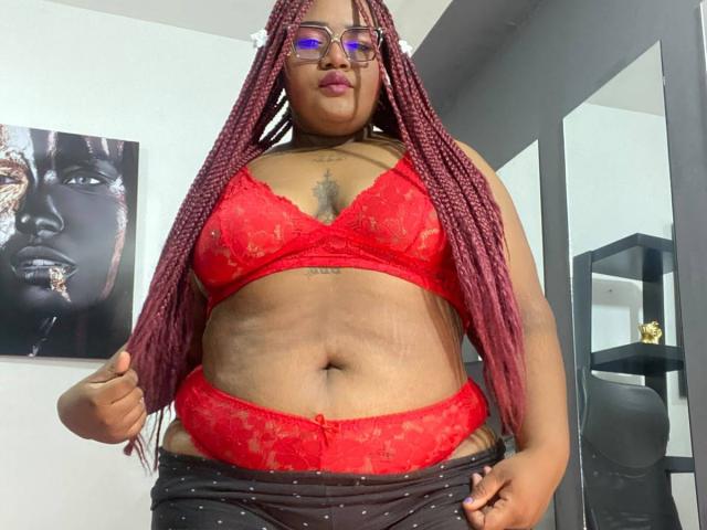 Bellamonroe, con un sujetador y tanga rojos de encaje, mostrando sus grandes senos y abdomen, tiene trenzas rojas largas, gafas y un tatuaje en el pecho, y está de pie en una habitación..