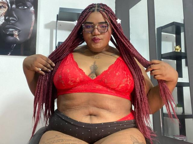 Bellamonroe mit roten Zöpfen, trägt einen roten Spitzen-BH und schwarze Punktehose, zeigt ihre Brüste und ihren Bauch, hält ihre Zöpfe, trägt eine Brille, Tattoos sind sichtbar, blickt in die Kamera..