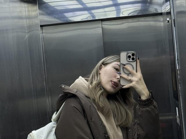 BebeNakaahiki with blonde hair, brown jacket, beige scarf, sticking out tongue, holding phone, mirror selfie, metallic background, white backpack, ring on finger, reflective surfaces.