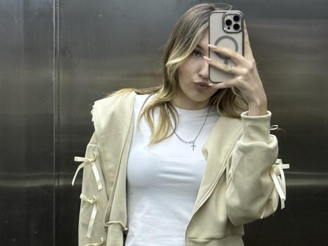 BebeNakaahiki met blond haar, een witte T-shirt, een beige jas met strikjes, die een telefoon vasthoudt, haar gezicht bedekt, een kruisje draagt, in een spiegel-selfie. Borsten zichtbaar onder het strakke T-shirt..