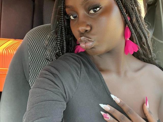 SexyDivanah-hot with dark skin, long braids, pink earrings, and glossy lips, wearing a gray top, showing her tits, and pink-nail-painted hand, sitting in a car.