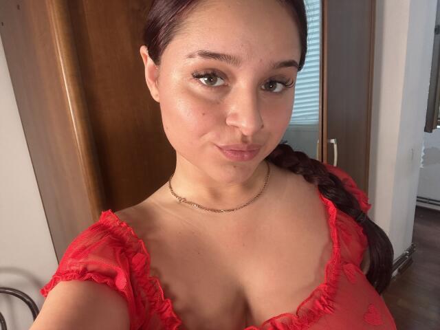 LucieRed has red braids, light brown skin, green eyes, and wears a red sheer top showing her tits. She's looking at the camera with a slight smile.