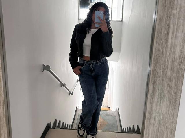 Sofia a les cheveux bouclés, porte une veste noire, un débardeur blanc et un jean bleu. Elle tient un téléphone, cachant son visage, et a les mains dans les poches..