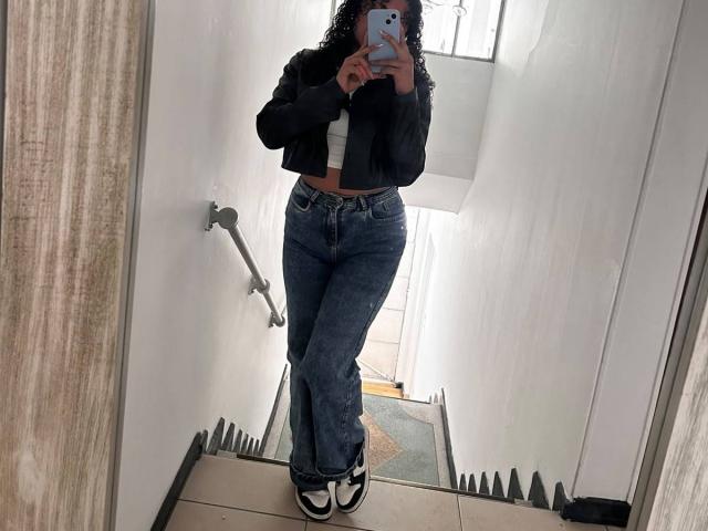 Sofia, passionnée, avec des cheveux bouclés, portant une veste noire, un crop top blanc, un jean bleu et des baskets noir et blanc, prenant un selfie devant un miroir dans un couloir blanc..