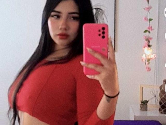 EmaFiore, con lunghi capelli neri, un top corto rosso e un telefono rosa, sta scattando un selfie allo specchio, mostrando il seno, tenendo il telefono nella mano destra, guardando la fotocamera, con un'espressione seria..