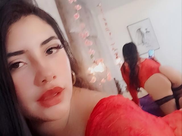 EmaFiore con rossetto rosso, sopracciglia scure e capelli neri, indossa una maglia rossa trasparente, e guarda uno specchio che riflette il suo sedere nudo e un laccio nero..