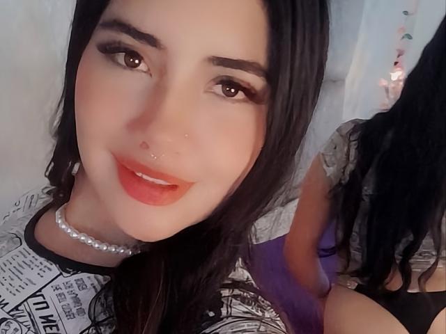 EmaFiore with dark hair, brown eyes, red lipstick, pearl necklace, and nose ring, smiling at the camera, showing her tits in a black and white patterned top.