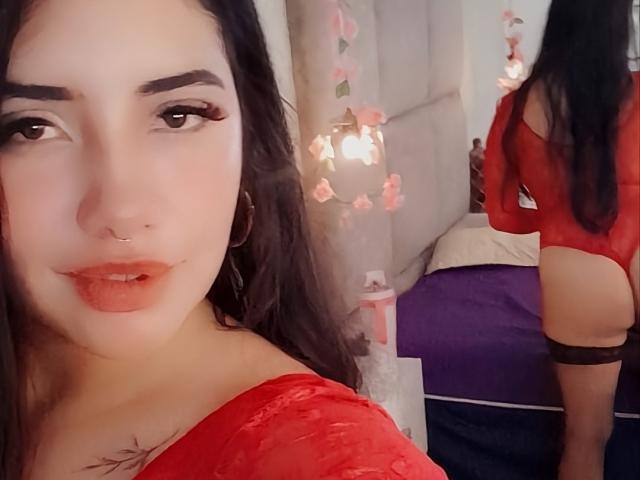 EmaFiore with red lace bodysuit, showing her ass in a mirror selfie. She has long black hair, dark eyebrows, red lipstick, and a nose ring. Reflection shows her back.