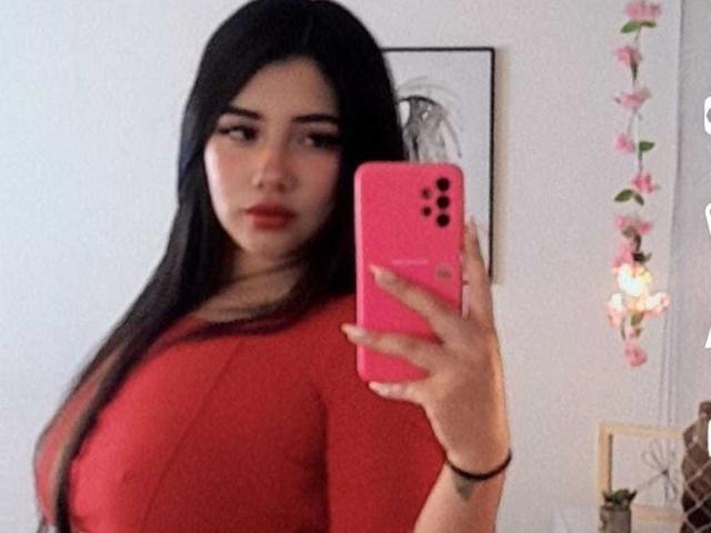 EmaFiore, con lunghi capelli neri, indossa una maglietta rossa, tiene in mano un telefono rosa, puntandolo verso lo specchio, mostrando il seno, guarda verso la fotocamera, con fiori rosa sullo sfondo, pelle chiara, labbra rosse..