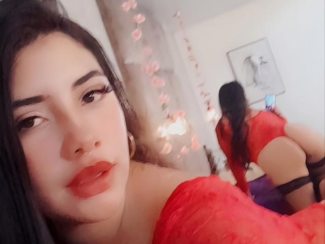 EmaFiore, avec ses cheveux foncés, son rouge à lèvres rouge et son haut en dentelle rouge, se montre dans un reflet de miroir, dévoilant ses fesses. Elle porte une jarretière noire et a un piercing au nez..