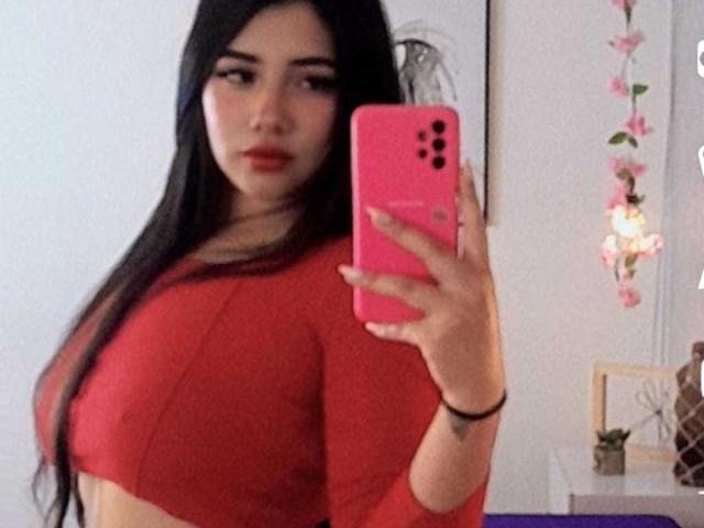 EmaFiore with long black hair, red crop top, and pink phone, taking a selfie, showing her big tits, holding phone in right hand, looking at camera with red lips.