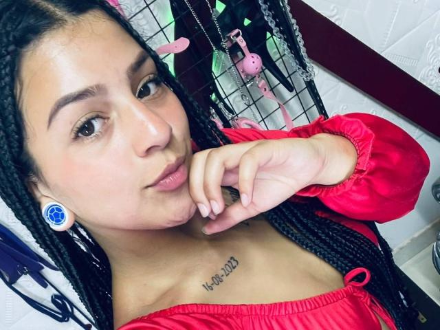 LonxEva with braided hair, red dress, blue earrings, brown eyes, and a tattoo under her tits that says "Kai" is pouting and touching her chin.