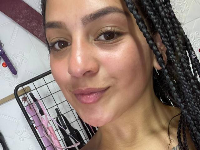LonxEva's face with braided black hair, brown eyes, and pink lipstick, smiling slightly with a smooth, slightly shiny complexion. Visible black ear gauges. Background includes a metal shelf.