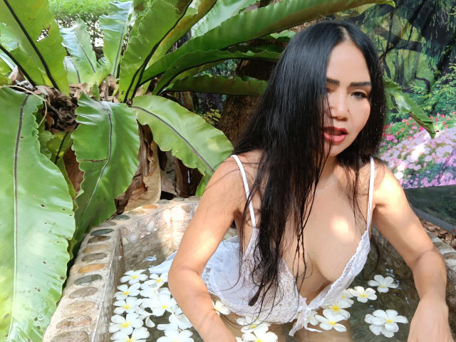 Lotusblume, con lunghi capelli neri, indossa una camicetta di pizzo bianco e ha il seno scoperto, è immersa in una vasca di pietra piena d'acqua e fiori bianchi di plumeria, è china in avanti con la bocca leggermente aperta..