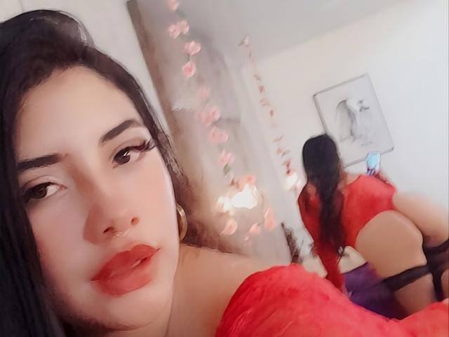 EmaFiore, com lábios vermelhos e cabelo preto, vestindo uma blusa vermelha com um ombro de fora, curvada, mostrando sua bunda nua, refletida em um espelho, com um efeito bokeh em forma de rosas..