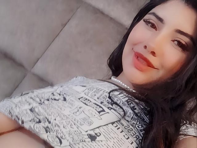 EmaFiore with long black hair, smiling, wearing a white graphic tee, pearl necklace, and red lipstick, lying on a gray couch, showing her tits slightly.
