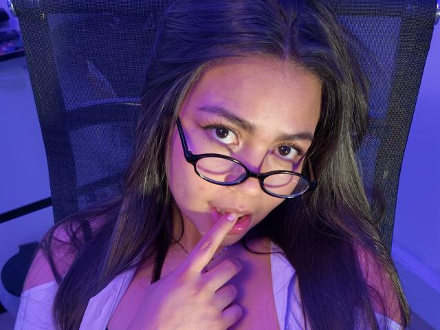 KimDevinne, com cabelos castanhos longos, usando óculos, mordendo o dedo, olhando para a câmera com iluminação roxo-azul, camisa branca, cadeira de malha preta, sexy, fofa, boca ligeiramente aberta..