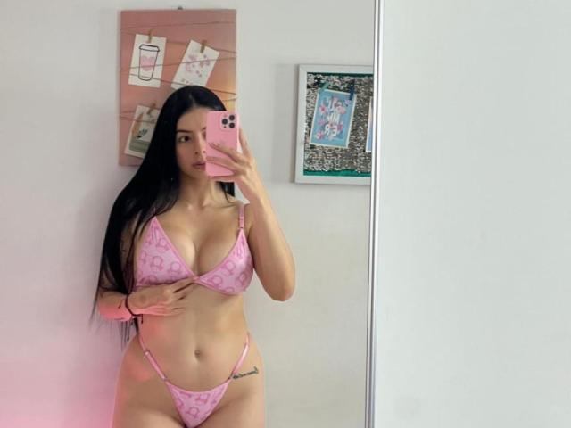 IsabellaJames with long black hair, pink patterned bikini, holding phone, showing her big tits and ass, right hand on her stomach, mirror selfie, tattoo on left hip.