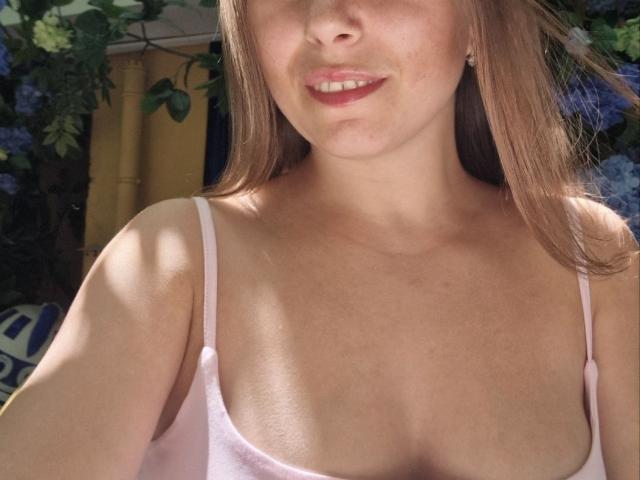 Jumiera's face with a subtle smile, pink spaghetti strap top showing small tits, light brown hair, fair skin, and red lipstick, surrounded by green and blue foliage.