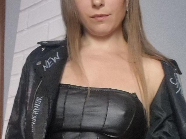 Jumiera with long blonde hair, wearing a black leather strapless top and a black leather jacket with white text, has small to medium tits and a slight smile.