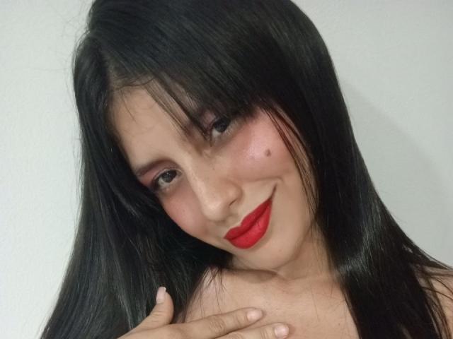 MadissonFontaine has long black hair, red lipstick, pink eyeshadow, and a small mole on her left cheek. She's smiling and touching her collarbone.