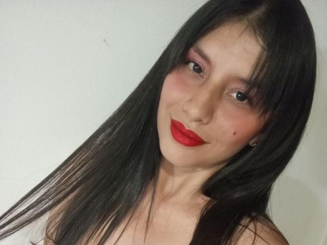MadissonFontaine has long black hair, red lipstick, pink eyeshadow, and a small mole on her right cheek, with a slight smile. She's wearing a white background.