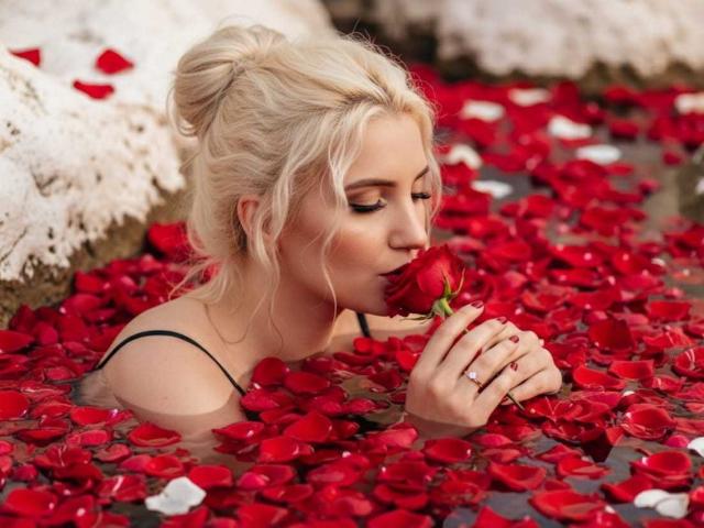 GiuliaAddams, blonde, dans une scène remplie d'eau avec des pétales de rose rouge, suce une rose rouge, sa main portant une bague est visible, et elle porte un haut noir à bretelles..