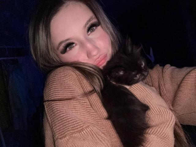 LatishaStreetman with long brown hair, wearing a tan ribbed sweater, holding a black cat with half-closed eyes against her cheek. Cat's head is resting on her chest.