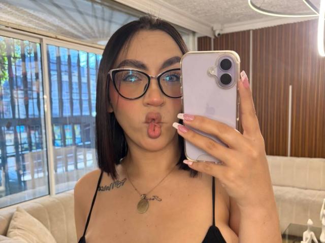 CamilleJuicyHot with glasses, pouty lips, black top, small tits, and a necklace, holding a white iPhone, taking a selfie in a room.