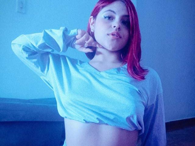 SophieHill with red hair, wearing a grey long-sleeve shirt, pulling it up to expose her midriff, looking at the camera with a slight pout.