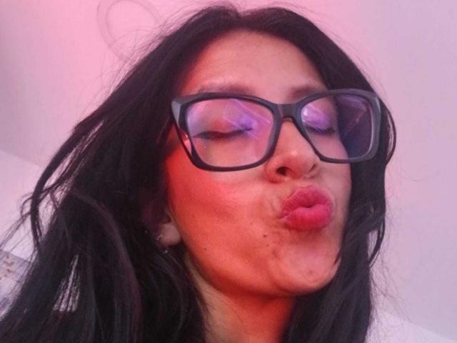 ZarayFeet, avec ses cheveux noirs, son rouge à lèvres rose vif et ses grandes lunettes noires, pouting ses lèvres pour un autoportrait. Son reflet montre un fard à paupières violet..