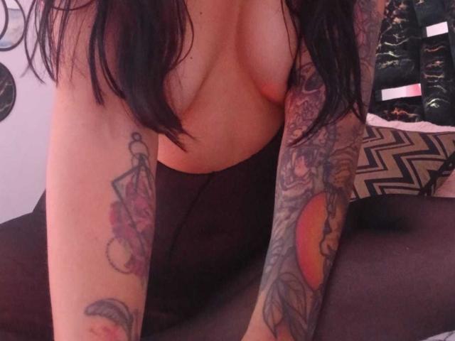 ZarayFeet, topless with dark hair, has tattoos on both arms, showing a pink diamond and orange sun, with black leggings and visible boobs. She's kneeling in a dim room.