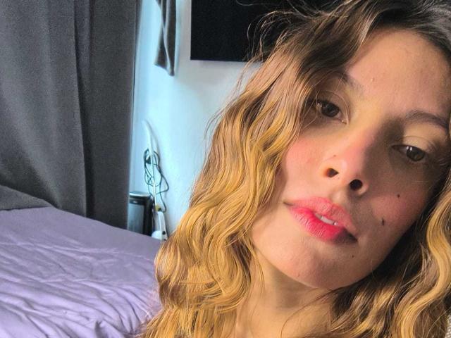 KiaraWiild tiene el cabello rubio ondulado, labios rosados y una pequeña marca de nacimiento en su mejilla derecha. Está mirando ligeramente hacia la izquierda con una sonrisa relajada y sutil..