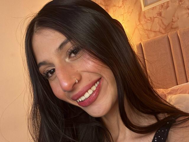 Sophie Beaumont tem cabelos pretos longos, um piercing no nariz e está sorrindo, usando batom rosa e com dentes brancos. Ela está vestindo uma blusa preta com bolinhas, e parece muito charmosa..