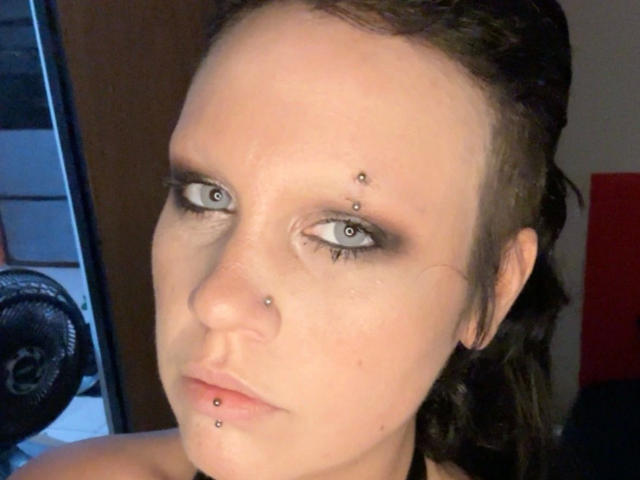 Kaotyka-hot hat Piercings in ihrer Nase und ihrem Unterlippe, sowie ein kleines Piercing oberhalb ihrer rechten Augenbraue. Sie hat blaue Augen, dunkles Haar und einen ernsten Gesichtsausdruck..