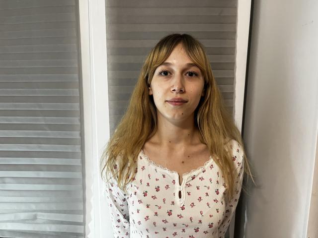 LanBordwell, con cabello rubio largo, senos pequeños, vistiendo una camisa blanca con estampado floral, de pie frente a persianas blancas, sonriendo ligeramente, piel clara, linda, informal..