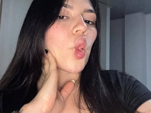 SophiaMarx with long black hair, pouty lips, and a hand touching her cheek, making a kissing face. She's wearing a black shirt, looking slightly bored.