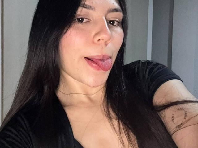 SophiaMarx has long black hair, slightly flushed cheeks, and is sticking her tongue out with a small tattoo on her upper right arm. She's wearing a black shirt.