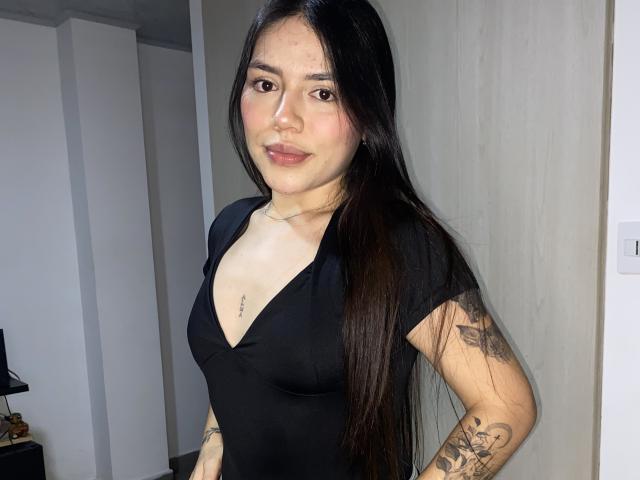 SophiaMarx with long black hair, wearing a black V-neck top, showing tattoos on her right arm and chest, standing in a room with a neutral background.