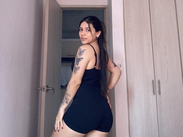 SophiaMarx stands in a doorway, wearing a black tank top and shorts, showing her ass and tattoos on her arms and side. She has long dark hair and looks back.