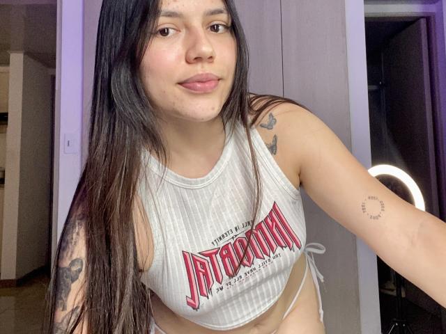 SophiaMarx with long dark hair, light skin, and tattoos, wearing a white ribbed tank top, showing her small tits and a ring tattoo on her right arm, smiling.