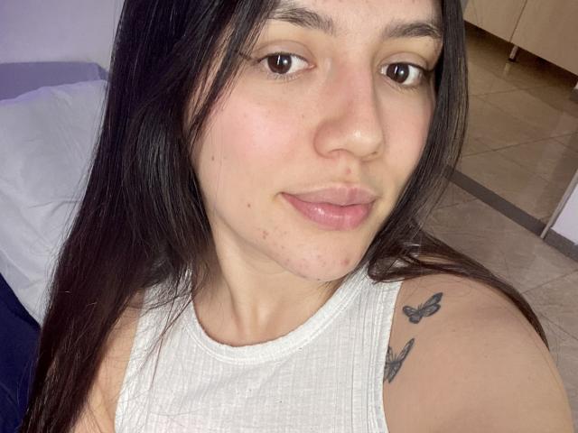 SophiaMarx has light skin, brown eyes, and long black hair. She's wearing a white tank top with butterfly tattoos on her shoulder. She's pouting with slightly glossy lips.