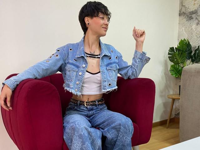 LissyLuxe está sentada em uma cadeira vermelha, vestindo uma jaqueta jeans azul com recortes, uma blusa cropped branca e calças jeans azuis combinando, olhando para o lado com um braço relaxado..