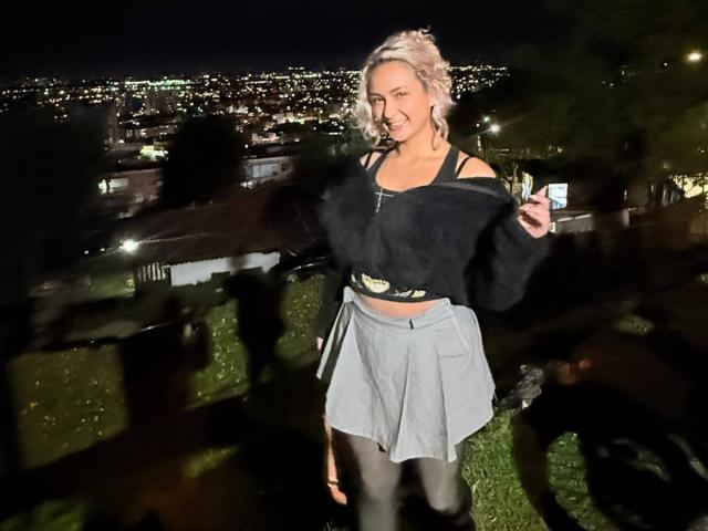 ElenaSaenz with blonde hair, wearing a black off-shoulder top and grey skirt, holding a phone, smiling, standing outdoors at night with city lights in the background.