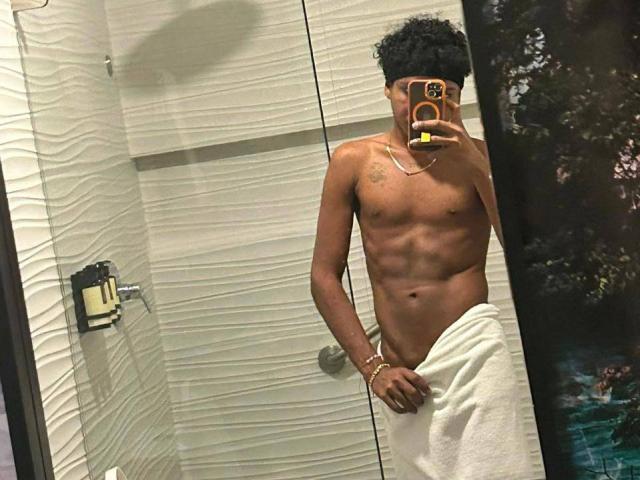 AlessandroBlack, sem camisa, com uma toalha branca cobrindo o traseiro, segurando um celular para tirar uma selfie no espelho, mostrando um torso musculoso e bronzeado, e cabelo preto encaracolado..