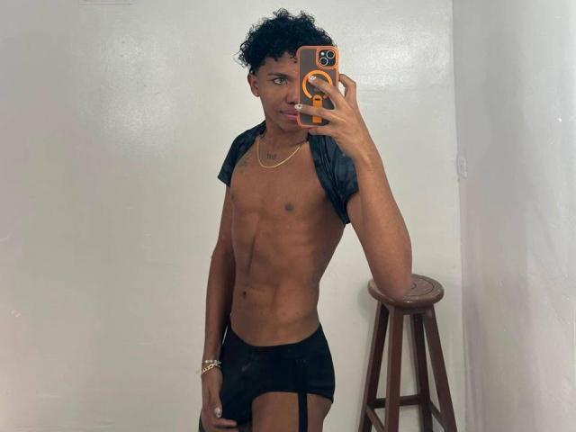 AlessandroBlack, con cabello negro rizado, vistiendo pantalones cortos negros y una camisa corta y abierta, sosteniendo un teléfono, mostrando un trasero musculoso, moreno y atractivo, y un collar de oro..