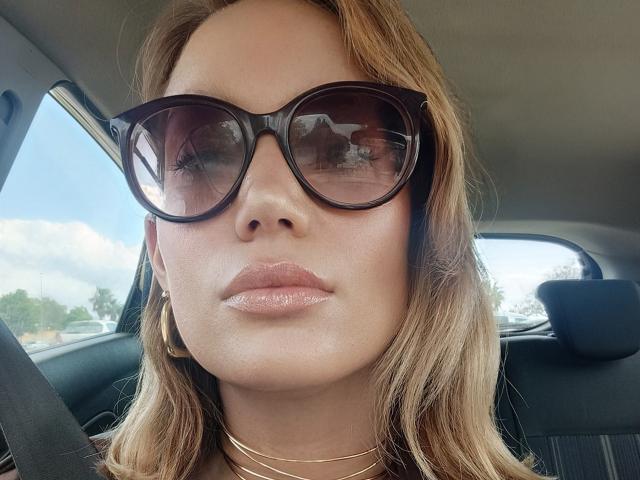 El rostro de MissFrenchy, con labios rosados y brillantes, lleva gafas de sol oscuras de gran tamaño, aros de oro y un collar de oro, sentada en un coche con cabello ondulado de color marrón claro..