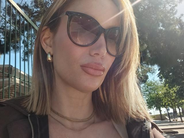 MissFrenchy, con labios rosados y sensuales, lleva gafas de sol negras, aretes dorados y una chaqueta negra, cabello rubio iluminado por el sol, un collar dorado, y está de pie al aire libre, con el sol brillando..