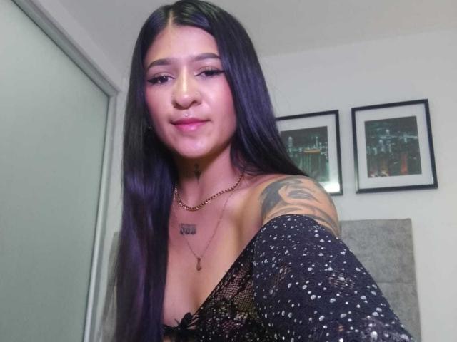 LuciadelaVega has long black hair, a nose piercing, and a tattoo on her right shoulder. She's wearing a black, speckled top with cleavage visible, and a necklace.